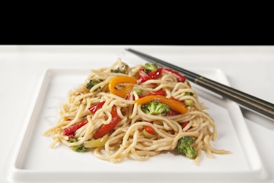 A plate of stir-fried noodles garnished with colorful vegetables such as broccoli, red and yellow bell peppers. The dish is presented on a white square plate with a pair of chopsticks resting beside it.