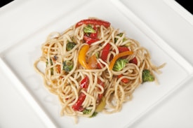 A plate of stir-fried noodles with a mix of colorful vegetables, including red and yellow bell peppers and broccoli florets, all lightly seasoned.