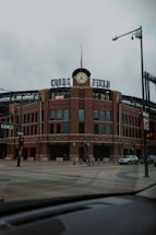A large brick sports stadium with a prominent clock tower and the words 'Coors Field' displayed on top. The street in the foreground is empty, with signage indicating Blake Street and various traffic lights visible.