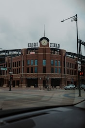A large brick sports stadium with a prominent clock tower and the words 'Coors Field' displayed on top. The street in the foreground is empty, with signage indicating Blake Street and various traffic lights visible.