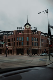 A large brick sports stadium with a prominent clock tower and the words 'Coors Field' displayed on top. The street in the foreground is empty, with signage indicating Blake Street and various traffic lights visible.
