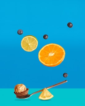 A floating arrangement of fruit slices including a lemon, an orange, and blueberries against a bright blue background. On the ground, a chocolate truffle in a wrapper is placed on a silver spoon alongside a lemon wedge.