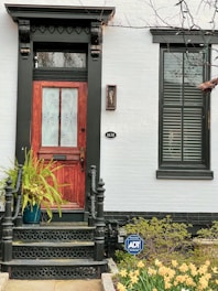 A wooden front door with intricate ironwork detail stands at the top of a small set of steps. The entryway is adorned with decorative black trim and features a potted fern on one side. The house number is mounted on the wall next to the door. A small ADT security sign is placed in the garden, which is filled with blooming yellow flowers and green shrubbery.