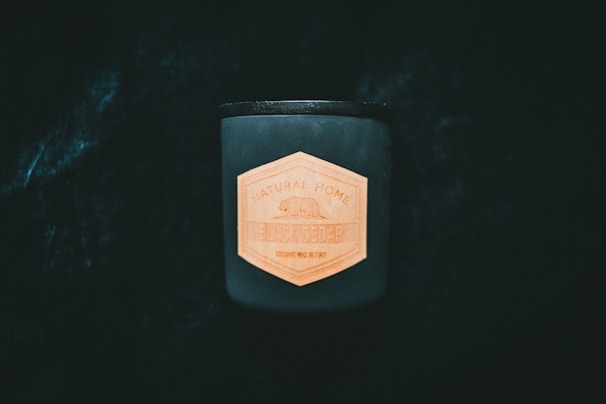 A dark, cylindrical candle stands out against a deep black background, featuring a hexagonal wooden label with the text 'Natural Home Black Cedar'. The label adds a natural, rustic touch to the modern design of the candle.