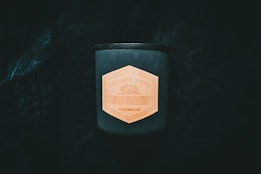 A dark, cylindrical candle stands out against a deep black background, featuring a hexagonal wooden label with the text 'Natural Home Black Cedar'. The label adds a natural, rustic touch to the modern design of the candle.