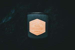 A dark, cylindrical candle stands out against a deep black background, featuring a hexagonal wooden label with the text 'Natural Home Black Cedar'. The label adds a natural, rustic touch to the modern design of the candle.