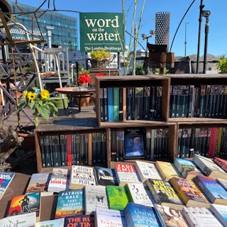 A variety of books displayed on shelves and a table, with a sign reading 'word on the water The London Bookbarge' in the background. The scene includes vibrant sunflowers and red flowers in pots, set against a bright blue sky.