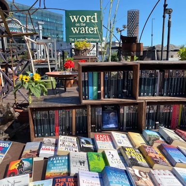 A variety of books displayed on shelves and a table, with a sign reading 'word on the water The London Bookbarge' in the background. The scene includes vibrant sunflowers and red flowers in pots, set against a bright blue sky.