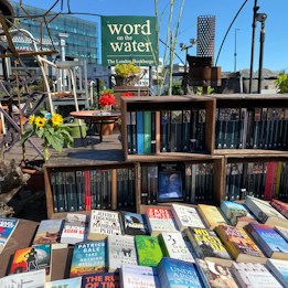 A variety of books displayed on shelves and a table, with a sign reading 'word on the water The London Bookbarge' in the background. The scene includes vibrant sunflowers and red flowers in pots, set against a bright blue sky.