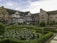 A historic, Tudor-style manor with intricate brickwork and wood paneling dominates the foreground. The building features large windows and elegant chimneys. In front of the manor, a formal garden with neatly trimmed hedges and symmetrical layouts enhances the estate's grandeur. The garden's meticulously arranged rows of rounded shrubs create a sense of order and elegance.