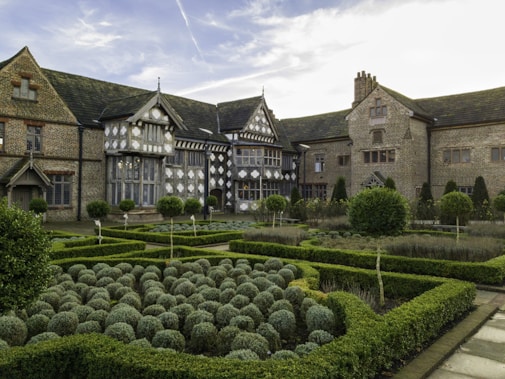 A historic, Tudor-style manor with intricate brickwork and wood paneling dominates the foreground. The building features large windows and elegant chimneys. In front of the manor, a formal garden with neatly trimmed hedges and symmetrical layouts enhances the estate's grandeur. The garden's meticulously arranged rows of rounded shrubs create a sense of order and elegance.