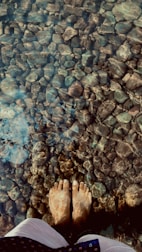 Bare feet standing in clear, shallow water over a bed of smooth stones, with light reflections creating patterns on the submerged rocks.