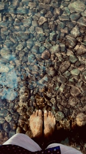 Bare feet standing in clear, shallow water over a bed of smooth stones, with light reflections creating patterns on the submerged rocks.