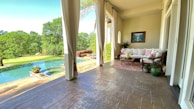 A serene outdoor patio features comfortable seating with white cushions and decorative pillows. The space is adorned with potted plants placed on a patterned rug. Large curtains frame the view outside, revealing a lush green landscape with trees and an infinity pool reflecting the sky.