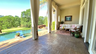 A serene outdoor patio features comfortable seating with white cushions and decorative pillows. The space is adorned with potted plants placed on a patterned rug. Large curtains frame the view outside, revealing a lush green landscape with trees and an infinity pool reflecting the sky.