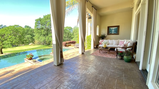 A serene outdoor patio features comfortable seating with white cushions and decorative pillows. The space is adorned with potted plants placed on a patterned rug. Large curtains frame the view outside, revealing a lush green landscape with trees and an infinity pool reflecting the sky.