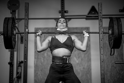 A person is lifting a barbell with heavy weights in a gym setting. The individual appears focused and is wearing a weightlifting belt and a wristwatch. The scene is captured in black and white, emphasizing the intensity and strength of the moment.