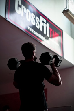 A person is holding two large dumbbells, standing under a sign that reads 'CrossFit' in a gym setting. The lighting is dim, casting a shadow on the individual who appears to be engaged in a workout.