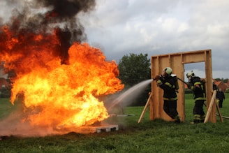 A group of firefighters in protective gear are actively extinguishing a large, intense fire with a powerful hose. The flames emit thick black smoke into the cloudy sky. The scene is set on a grassy field with a wooden frame structure nearby.