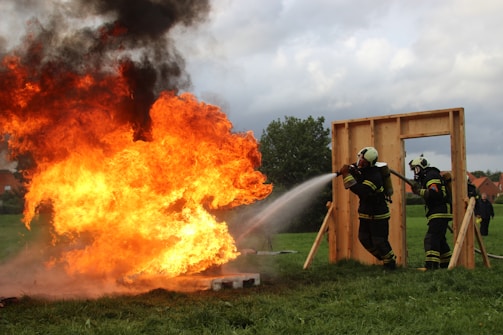 A group of firefighters in protective gear are actively extinguishing a large, intense fire with a powerful hose. The flames emit thick black smoke into the cloudy sky. The scene is set on a grassy field with a wooden frame structure nearby.