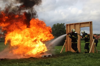 A group of firefighters in protective gear are actively extinguishing a large, intense fire with a powerful hose. The flames emit thick black smoke into the cloudy sky. The scene is set on a grassy field with a wooden frame structure nearby.