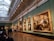 An art gallery with high arched ceilings showcases large classical paintings in ornate gold frames on green walls. People are viewing the artworks, with some sitting on benches and others standing.
