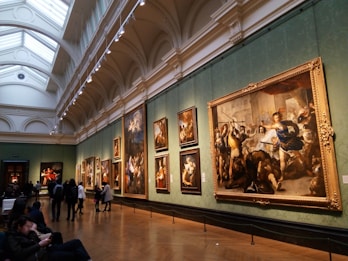 An art gallery with high arched ceilings showcases large classical paintings in ornate gold frames on green walls. People are viewing the artworks, with some sitting on benches and others standing.