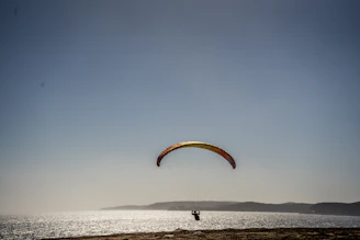A person is paragliding over a scenic coastal area with calm waters and distant hills. The paraglider's canopy is colorful, and the clear sky enhances the expansive view.
