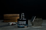 A bottle of Yves Saint Laurent L'Homme Le Parfum is placed on a surface, with its cap off and positioned to the right. There is a wristwatch laid down to the left of the bottle and a wooden box in the background.