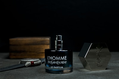 A bottle of Yves Saint Laurent L'Homme Le Parfum is placed on a surface, with its cap off and positioned to the right. There is a wristwatch laid down to the left of the bottle and a wooden box in the background.