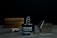 A bottle of Yves Saint Laurent L'Homme Le Parfum is placed on a surface, with its cap off and positioned to the right. There is a wristwatch laid down to the left of the bottle and a wooden box in the background.