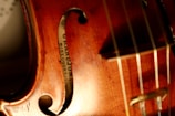 Close-up view of a violin showcasing its warm wooden texture with visible wood grain. The image captures part of the violin's body, F-hole, strings, and a label inside which reads 'GRANDJON MIRECOURT'. The lighting highlights the instrument's glossy finish and fine craftsmanship.