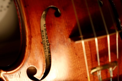 Close-up view of a violin showcasing its warm wooden texture with visible wood grain. The image captures part of the violin's body, F-hole, strings, and a label inside which reads 'GRANDJON MIRECOURT'. The lighting highlights the instrument's glossy finish and fine craftsmanship.