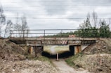 A small concrete bridge with graffiti of the word 'Canada' is situated over a narrow dirt path. The bridge has rusty metal railings and is surrounded by sparse greenery. Overhead, power lines stretch across the sky. The landscape is overcast, with a mix of bare trees and coniferous trees.