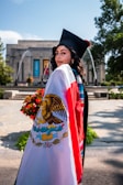 A graduate in a cap and gown is standing in an outdoor setting, draped in a Mexican flag. The background features a building with columns and colorful banners, as well as fountains and greenery.