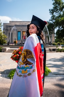 A graduate in a cap and gown is standing in an outdoor setting, draped in a Mexican flag. The background features a building with columns and colorful banners, as well as fountains and greenery.