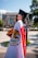 A graduate in a cap and gown is standing in an outdoor setting, draped in a Mexican flag. The background features a building with columns and colorful banners, as well as fountains and greenery.