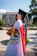 A graduate in a cap and gown is standing in an outdoor setting, draped in a Mexican flag. The background features a building with columns and colorful banners, as well as fountains and greenery.