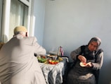 An elderly woman and man are sitting on a balcony, with a table covered in a patterned cloth between them. The woman is preparing food, holding an open sandwich with vegetables, while the man is facing away. Various dishes, vegetables, and a teapot are on the table, and sunlight streams in through a window.