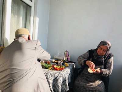 An elderly woman and man are sitting on a balcony, with a table covered in a patterned cloth between them. The woman is preparing food, holding an open sandwich with vegetables, while the man is facing away. Various dishes, vegetables, and a teapot are on the table, and sunlight streams in through a window.