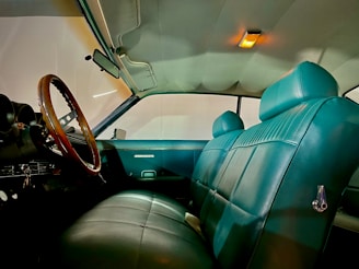 The interior of a vintage car featuring teal leather seats and a wood-grain steering wheel with a metal trim. The ceiling light casts a warm glow, highlighting the spacious and well-maintained seating area.