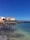 A vibrant seaside scene with several people enjoying a beach. The water is clear with a mix of turquoise and darker shades. Behind the beach, there is a promenade lined with a variety of buildings in colors such as orange, white, and beige. The sky is clear and deep blue.