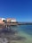 A vibrant seaside scene with several people enjoying a beach. The water is clear with a mix of turquoise and darker shades. Behind the beach, there is a promenade lined with a variety of buildings in colors such as orange, white, and beige. The sky is clear and deep blue.