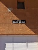 A tan-colored brick wall with a small, square window near the top. Below the window, there is a dark rectangular sign with white text reading 'EDIFICIO BALCON DEL MAR.' The lower portion of the image shows a white shuttered window or door.