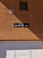 A tan-colored brick wall with a small, square window near the top. Below the window, there is a dark rectangular sign with white text reading 'EDIFICIO BALCON DEL MAR.' The lower portion of the image shows a white shuttered window or door.