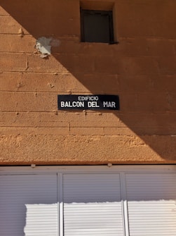 A tan-colored brick wall with a small, square window near the top. Below the window, there is a dark rectangular sign with white text reading 'EDIFICIO BALCON DEL MAR.' The lower portion of the image shows a white shuttered window or door.