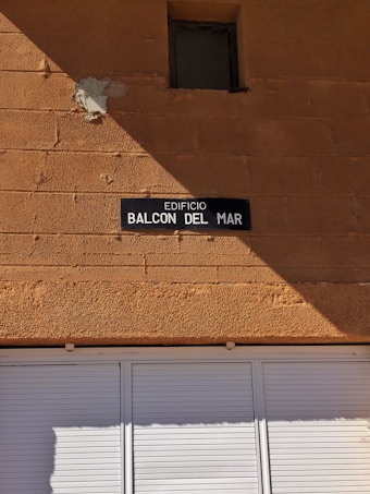 A tan-colored brick wall with a small, square window near the top. Below the window, there is a dark rectangular sign with white text reading 'EDIFICIO BALCON DEL MAR.' The lower portion of the image shows a white shuttered window or door.