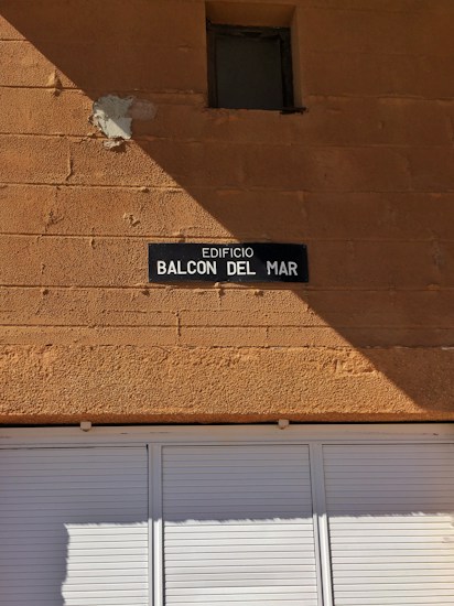 A tan-colored brick wall with a small, square window near the top. Below the window, there is a dark rectangular sign with white text reading 'EDIFICIO BALCON DEL MAR.' The lower portion of the image shows a white shuttered window or door.