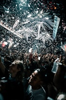 A lively crowd is celebrating under a massive indoor installation with bright, decorative elements hanging from the ceiling. Confetti is falling everywhere, and people are cheering and holding up their phones, capturing the moment.