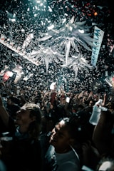 A lively crowd is celebrating under a massive indoor installation with bright, decorative elements hanging from the ceiling. Confetti is falling everywhere, and people are cheering and holding up their phones, capturing the moment.